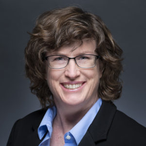 Headshot shows a White woman with an open smile, wearing glasses with thin black frames. Her hair is medium-length and wavy-brown. Her eyes are smiling. The collar of her shirt is light blue and her suit jacket is black.